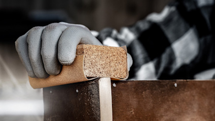 En hand i grå handske slipar en träram med ett sandblock, bär rutigt skjorta i bakgrunden.