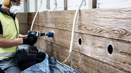 En person borrar hål i en träpanel med en sladdlös borrmaskin, skyddsutrustning används.