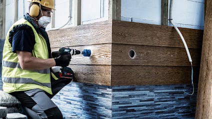 Person i skyddshjälm och väst använder borrmaskin vid panel av trä och kakel.