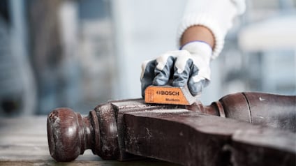 Person slipar ett mörkt trämöbelben med ett Bosch-slipblock.