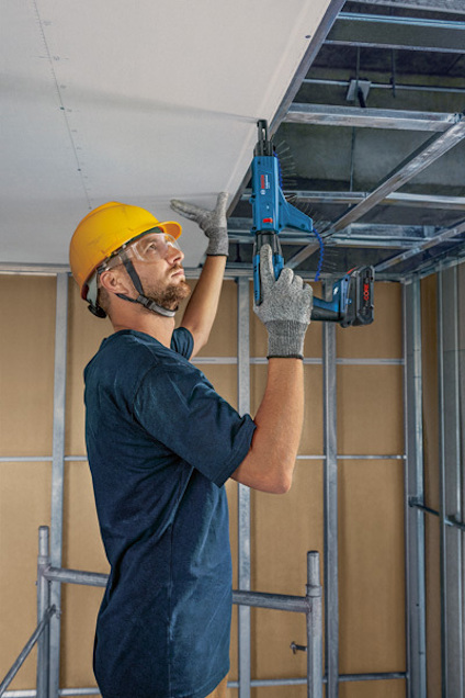 Person med gul hjälm och skyddsglasögon skruvar i tak med elverktyg, omgiven av väggar av gipsskivor och metallreglar.