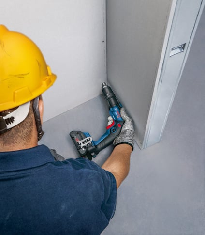 Person med gul hjälm och handskar använder en skruvdragare för att fästa en gipsskiva.