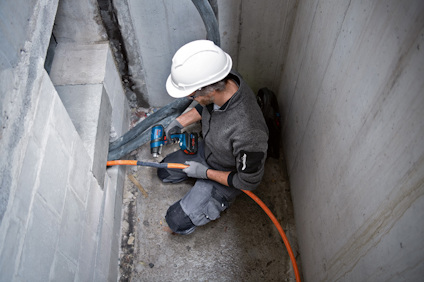 Person i arbetskläder borrar i betongvägg med borrmaskin, iförd vit hjälm och skyddshandskar.
