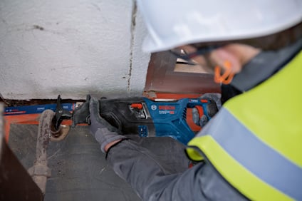 Person med hjälm och skyddsväst använder elektrisk såg mot en vägg.