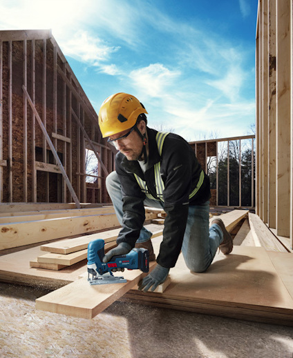 Person i hjälm och skyddsglasögon sågar träplankor på en byggarbetsplats.