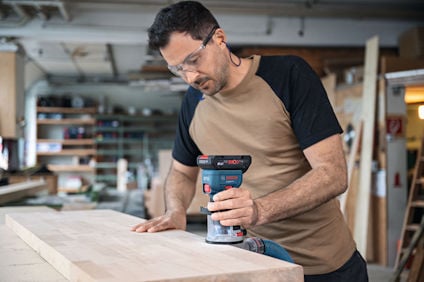 Person använder slipmaskin på träskiva i verkstad. Bär skyddsbrillor.