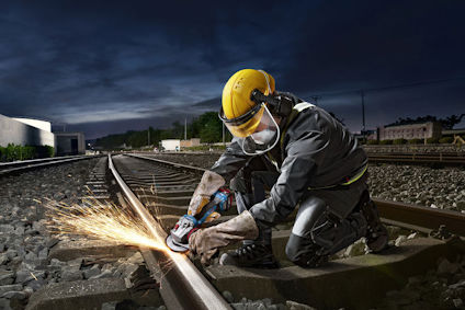 Person med skyddsutrustning slipar järnvägsräls med gnistor i mörk miljö.