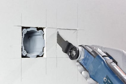 En oscil­lerande såg skär ett fyrkantigt hål i en gipsvägg, bredvid ett insatt vägguttag.