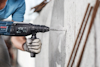 Person i blå tröja och skyddshandskar borrar i betongvägg med Bosch-borrmaskin.
