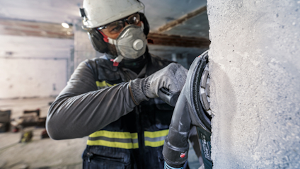 Person med skyddsutrustning använder en elverktyg för att slipa betongyta i en byggnad.