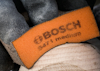 Arbetsvante med orange läderdel och texten "BOSCH ser i medium", handen håller något träaktigt.