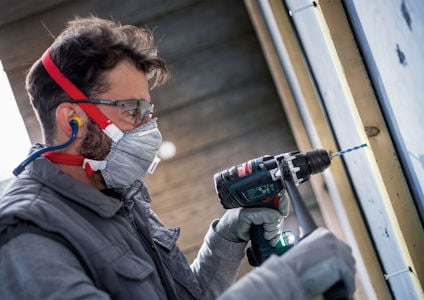 En person borrar med en borrmaskin och bär skyddsutrustning som hörselskydd, skyddsglasögon och ansiktsmask.