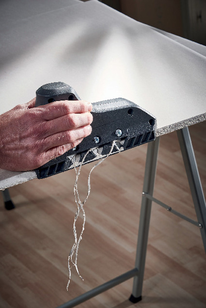 En person använder en handhållen skärare för att dela en gipsskiva, synliga trådar lossnar från skåran.