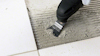 En person använder en platt spackelspade för att applicera eller jämna ut grått murbruk på kakelunderlag.